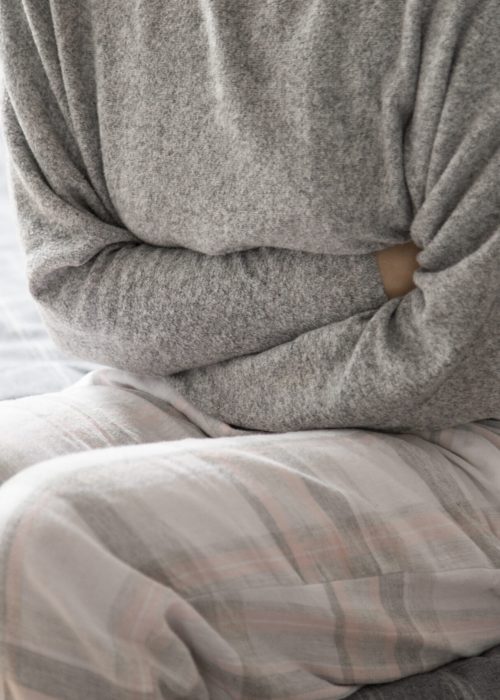 Sick woman in grey homewear sitting on bed, keeping hands on stomach, suffering from pain. Illness, stomach ache concept