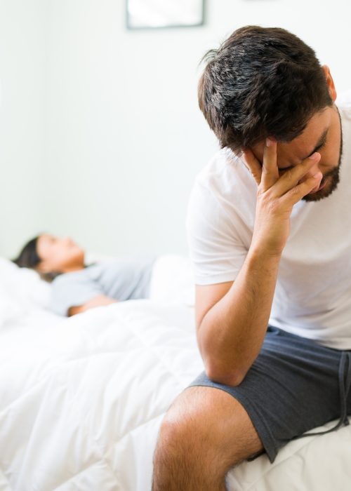 Thoughtful young man in pjs sitting up in bed in the morning and feeling sad with his bad relationship